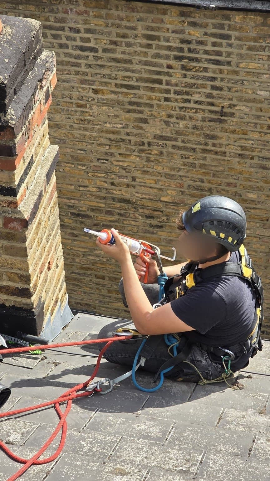 Rope access technician applying sealant by gun at the base of a brick chimney stack